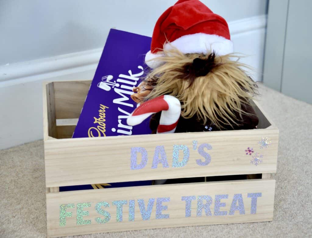 wooden crate with a soft toy and large chocolate bar inside. The front of the crate has sparkly lettering cut with a Cricut Joy saying 'Dad's Festive Treats'